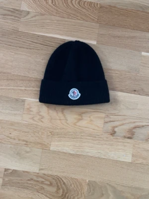 Svart Moncler mössa med logotyp - Säljer en svart ribbstickad mössa från Moncler med uppvikt kant och klassisk Moncler-logotyp framtill. Mössan är enkel och stilren, perfekt för dig som vill ha en snygg accessoar med exklusiv känsla. Materialet är mjukt och värmande.
