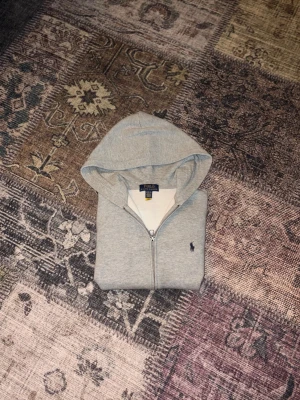 Grå hoodie från Polo Ralph Lauren - Snygg grå hoodie med dragkedja från Polo Ralph Lauren. Klassisk design med huva, fickor framtill och den ikoniska lilla broderade loggan på bröstet. Perfekt för en avslappnad och stilren look.