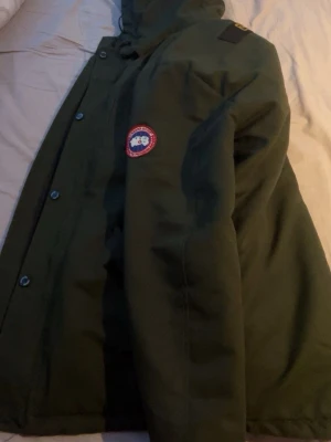 Mörkgrön parkas från Canada Goose - Säljer en mörkgrön parkas från Canada Goose med klassisk logotyp på ärmen. Jackan har huva, tryckknappar och dragkedja framtill samt två stora fickor. Perfekt för kalla dagar och riktigt snygg streetstil. Storlek L men passar även M