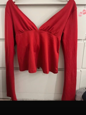 Röd långärmad topp med knyt - Trendig röd topp med djup v-ringning och långa ärmar. Toppen har en snygg knyt-detalj i ryggen som ger en unik look. Tillverkad i mjukt bomullsmaterial och har en lätt utsvängd passform nertill. Perfekt för dig som vill sticka ut.