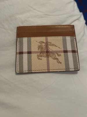 Burberry kortfodral i brunt läder - Snyggt kortfodral från Burberry i brunt läder och klassiskt rutigt tyg med ridmotiv. Fodralet har flera kortfack och den ikoniska Burberry-loggan på framsidan. Perfekt för dig som vill ha en stilren och lyxig accessoar i fickan. ///Priset är diskuterbart!!!