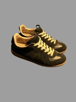 Masion margelas gats  - Snygga svarta sneakers med beige detaljer och klassisk gummisula. Skorna har vita snören och en låg silhuett. Materialet är en mix av mocka och läder för en stilren look. Perfekta för dig som gillar minimalistisk och tidlös design.