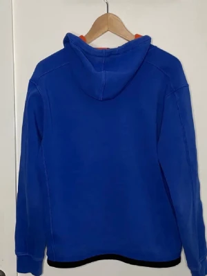 Blå hoodie från Polo Ralph Lauren - Snygg blå hoodie från Polo Ralph Lauren med dragkedja framtill och två svarta fickor med dragkedja. Insidan av huvan är orange och bröstet pryds av den klassiska Polo-loggan i orange. Perfekt för en chill och sportig look. Dm för fler bilder.