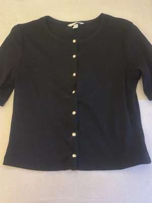 Svart ribbad kofta med pärlknappar - Stilren svart ribbad kofta med rund halsringning och pärlknappar framtill. Modellen är croppad och har trekvartsärmar. Perfekt att slänga över en topp för en snygg lager-på-lager-look.