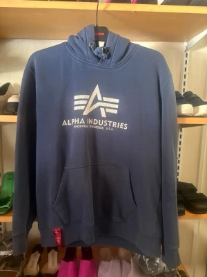 Blå hoodie från Alpha Industries - Snygg blå hoodie från Alpha Industries med vit logga och text framtill. Klassisk känguruficka och justerbar huva med dragsko. Mjuk bomullskänsla och ribbade muddar vid ärmslut och nederkant. Perfekt för en avslappnad streetstyle-look.