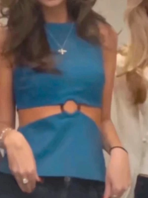 Blå croppad topp med cut out och ring - Snygg blå croppad topp med cut out-detaljer i midjan och en dekorativ ring framtill. Toppen är ärmlös och har en figurnära passform som framhäver midjan. Perfekt för dig som vill sticka ut lite extra.