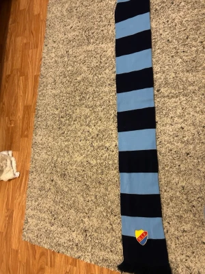 Randig Djurgården halsduk blå/svart - Snygg halsduk med breda blå och svarta ränder från Djurgårdens IF. Halsduken har DIF:s klubbmärke broderat i ena änden och fransar längst ner. Perfekt för att visa ditt stöd på matchen eller i vardagen.