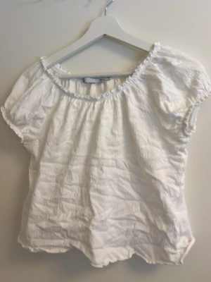 Vit blus med resårdetaljer - Söt vit blus med korta ärmar och resår vid halsringning och nederkant. Tillverkad i ett lätt, skrynkligt bomullstyg som ger en avslappnad känsla. Perfekt till jeans eller kjol för en enkel och fräsch look.