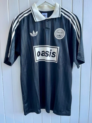 Oasis - Turné 25 - Fotbollströja (L) - Säljer en svart Oasis-supportertröja för bandets turné under 2025. Tröjan är en reprint i nyskick. Aldrig använd. ”25” på ryggen.    Storlek: Large  #Kent #Cd #Vinyl #England