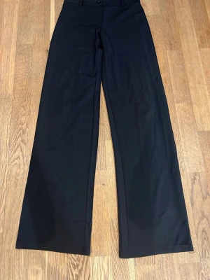 Svarta vida kostymbyxor från Nelly - Snygga svarta kostymbyxor från Nelly med vida ben och hög midja. Byxorna har klassisk design med knapp och dragkedja framtill.  Helt oanvända då de inte passade