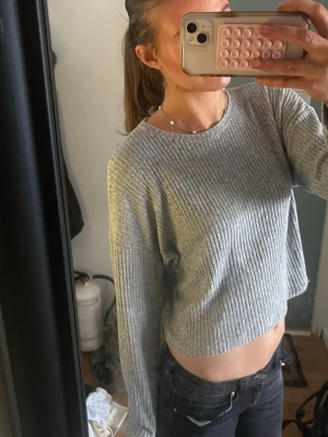 Grå ribbstickad cropped tröja - Säljer en grå ribbstickad cropped tröja med rund hals och långa ärmar. Tröjan har en enkel och stilren design, perfekt att matcha med jeans eller kjol. Materialet är mjukt och bekvämt, och modellen är lite lös för en avslappnad look.