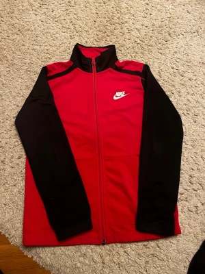Röd och svart zip-jacka från Nike stl 140 (barn)  - Snygg sportjacka från Nike i rött med svarta ärmar och axelparti. Jackan har hel dragkedja framtill, ståkrage och vit Nike-logga på bröstet. Perfekt för träning eller chill. Standard fit och tillverkad i mjukt material.
