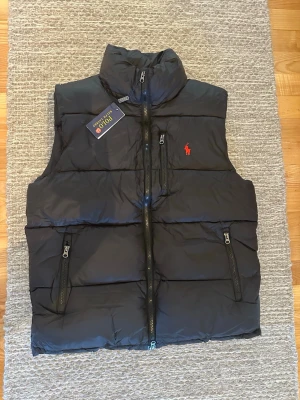 Svart dunväst från Polo Ralph Lauren - Snygg svart dunväst från Polo Ralph Lauren med hög krage och dragkedja framtill. Västen har två sidofickor med dragkedja och en bröstficka. Klassisk röd logga broderad på bröstet. Perfekt för lager på lager och streetwear-stil. STORLEK M