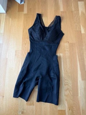 Svart shapingbody med spetsdetaljer - Snygg svart shapingbody med breda axelband och v-ringad hals. Överdelen har fina spetsdetaljer och mesh, medan nederdelen är slät och formande med ben som går till mitten av låret. Perfekt för att skapa en smickrande siluett under kläderna.