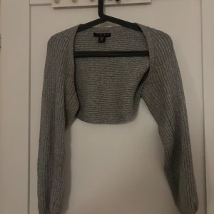 Grå ribbstickad bolero från Gina Tricot - Kort grå bolero från Gina Tricot i ribbstickat material. Långärmad modell med öppen front, perfekt att slänga över en topp eller klänning. Enkel och stilren design som passar till många outfits.