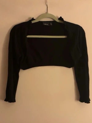 Svart bolero med volangkant - Kort svart bolero med trekvarts ärmar och volangkant längs öppningen. 