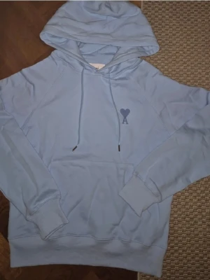 Ljusblå hoodie från Ami Paris - Säljer en ljusblå hoodie från Ami Paris med klassisk huva och dragsnören. På bröstet finns det ikoniska hjärta-A broderiet. Hoodien har känguruficka och ribbade muddar. Perfekt för en chill och stilren look.