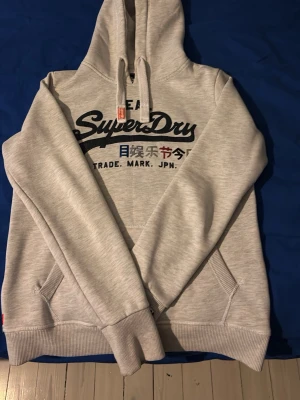 Ljusgrå hoodie från Superdry - Snygg ljusgrå hoodie från Superdry med svart och färgglad logga framtill samt japanska tecken. Klassisk modell med huva och dragsko, samt känguruficka. Perfekt för en chill och avslappnad stil.