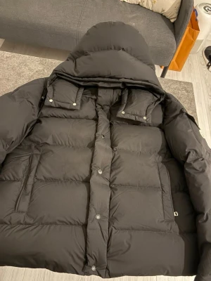 Svart pufferjacka med huva - Snygg svart pufferjacka med stor huva och två bröstfickor med lock och knappar. Jackan har även två sidofickor och stängs med tryckknappar framtill. Perfekt för kalla vinterdagar tack vare det tjocka, vadderade materialet.