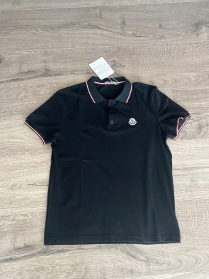 Svart piké från Moncler med röd/vita detaljer - Snygg svart pikétröja från Moncler med klassisk krage och knappar framtill. Röd och vit rand längs krage och ärmslut ger en sportig touch. Ikonisk Moncler-logga på bröstet. Tillverkad i mjuk bomull för skön känsla.