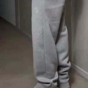 Gråa mjukisbyxor från suvene flared - Säljer ett par stilrena gråa mjukisbyxor från suvene med broderad logga på benet. Byxorna har avslappnad passform, resår i midjan och muddar vid bensluten. Perfekta för chill dagar eller när du vill ha det bekvämt.