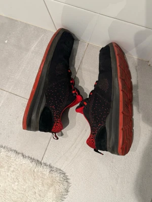 Svarta och röda sneakers med mesh - Säljer ett par svarta sneakers med röda detaljer och röd sula. Skorna har mesh-överdel för bra ventilation och svarta partier i mockaimitation vid hälen. Snygga röda skosnören och prickiga detaljer längs sidorna ger en sportig look.