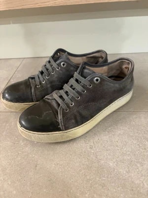 Lanvins skor uk 5 - Säljer ett par svarta låga sneakers med ovandel i canvas och tåparti i blankt läder. Skorna har vita sulor och grå skosnören. Perfekta för en avslappnad streetstil och passar till det mesta i garderoben.