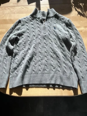 Grå kabelstickad tröja Polo Ralph Lauren - Kabelstickad grå tröja från Polo Ralph Lauren med halv dragkedja och broderad logga på bröstet. Tröjan har ribbade muddar och krage, samt klassisk kabelstickad struktur. Perfekt för lager-på-lager och chill stil.