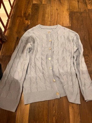 Ljusblå kabelstickad kofta - Superfin ljusblå kofta med kabelstickat mönster och snygga knappar framtill. Koftan har rund halsringning, långa ärmar och ribbade muddar. Perfekt att slänga på sig när det är lite kyligt ute.