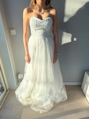 Vit långklänning/bröllopsklänning  - Elegant vit långklänning med axelbandslös design och vacker pärlbroderad överdel. Kjolen är i flera lager av tunt, svepande tyg som ger en drömmig känsla. Perfekt bröllops klänning eller för en bal! Nypris 3999, klänningen är helt ny i storlek 34 från bubbleroom. Man kan välja att antingen köra utan de axelbanden eller bara knäppa på dom. Klänningen har även en liten slits innanför tyget som syns när man går. Skriv privat för fler bilder eller funderingar. 