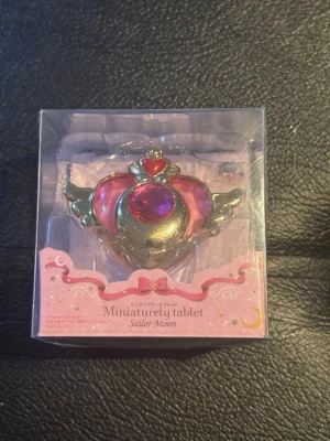 Sailor Moon hjärtformad brosch - Hjärtformad brosch från Sailor Moon i metall-look med rosa och röda detaljer samt vingar på sidorna. Broschen har en stor röd sten i mitten och små dekorativa hjärtan. Perfekt för cosplay eller maskerad och kommer i originalförpackning.