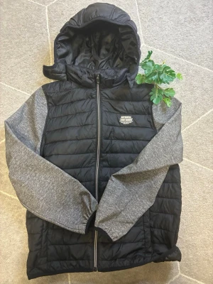 Svart pufferjacka med grå ärmar - Snygg svart pufferjacka från Authentic Apparel med gråmelerade ärmar och huva. Jackan har quiltad design, dragkedja framtill och en liten logga på bröstet. Perfekt för kalla dagar och riktigt skön att bära.