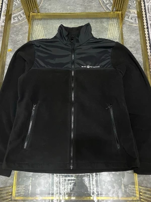 Svart fleecejacka från Sail Racing - Svart fleecejacka från Sail Racing med hög krage och dragkedja framtill. Jackan har två sidofickor med dragkedja och detaljer i blankt syntetmaterial över bröstet. Perfekt för kyliga dagar och enkel att matcha med allt.