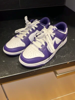 Nike Dunk Low vit/lila sneakers - Snygga Nike Dunk Low sneakers i vitt och lila läder med klassisk swoosh på sidorna. Skorna har vita skosnören, perforerad tåbox och lila detaljer runt häl och sula. Perfekt för dig som vill sticka ut med en ikonisk och trendig modell. Använd 1-2 gånger så väldigt bra skick.