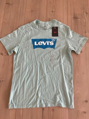 Ljusblå Levi's t-shirt med logga - Snygg ljusblå t-shirt från Levi's med stor blå och vit logga på bröstet. Klassisk rund hals och korta ärmar. Tillverkad i mjuk bomull, perfekt för en avslappnad stil.