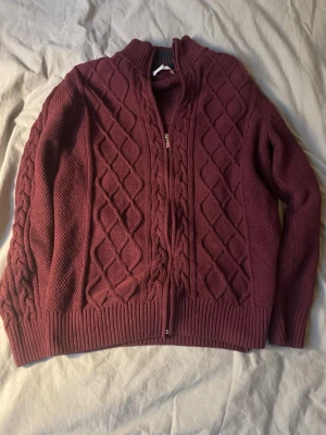Vinröd kabelstickad ziptröja - Mysig vinröd stickad tröja med dragkedja framtill och kabelstickat mönster. Tröjan har hög krage, ribbade muddar och är tillverkad i ett tjockare garn som passar perfekt när det är kallt ute.