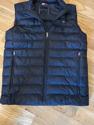 Mörkblå dunväst från Tommy Hilfiger strl S - Snygg mörkblå dunväst från Tommy Hilfiger Storlek S med quiltad design och två dragkedjefickor framtill. Västen har hög krage och diskret logga på bröstet. Perfekt för lager-på-lager och ger en stilren look till din outfit.