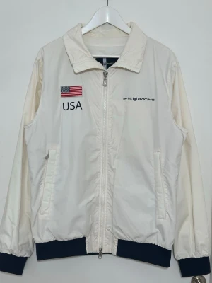 Vit vindjacka Sail Racing USA - Vit vindjacka från Sail Racing med broderad USA-flagga och texten 'USA' fram samt 'UNITED STATES' och flagga bak. Jackan har dragkedja, två fickor och mörkblå muddar vid ärmslut och nederkant. 