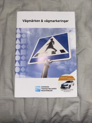 Vägmärken & vägmarkeringar bok - Kurslitteratur med titeln 'Vägmärken & vägmarkeringar' från Sveriges Trafikutbildares Riksförbund. Omslaget är vitt och blått med en bild på ett övergångsställe och STR-logga. Perfekt för dig som pluggar till körkort.