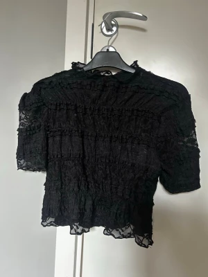 Svart spetsblus med volangdetaljer - Trendig svart blus i genombruten spets med volangdetaljer och korta ärmar. Croppad modell som ger en cool och edgy vibe. Perfekt att styla med höga jeans eller kjol för en snygg look.