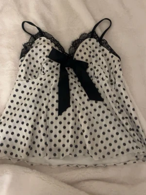Prickigt linne med spets och rosett - Supersött vitt linne med svarta prickar, tunna axelband och dekorativ svart spets vid bysten. Stor svart rosett framtill ger en extra gullig touch. Luftigt och bekvämt material, perfekt för mysiga kvällar. 