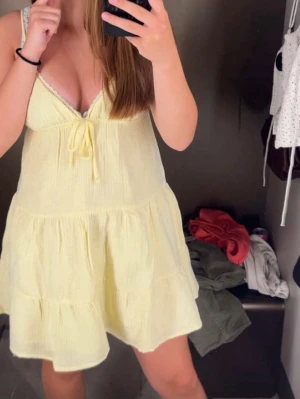 Ljusgul kort sommarklänning - Söt ljusgul kort klänning med v-ringning och knyt framtill. Klänningen har volangdetaljer och är ärmlös, perfekt för varma dagar. Tillverkad i ett lätt och luftigt bomullstyg som ger en avslappnad och somrig känsla.