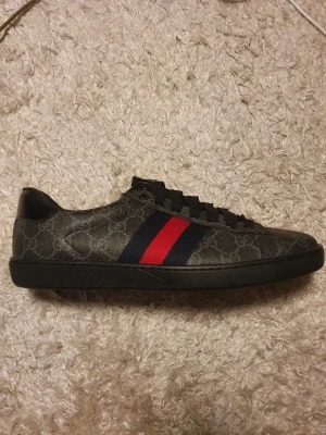 Gucci Ace sneakers med GG-mönster - Oanvända, endasr gått med inomhus.    Svarta Gucci Ace sneakers med klassiskt GG-monogram i canvas, blå och röda ränder på sidan samt metallic-detaljer i rött och blått på hälen. Snygg och lyxig design med läderdetaljer och tydlig logga på baksidan. Kommer med originalkartong och dustbag.