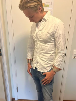 Vit burberry skjorta  - Säljer en vit skjorta med klassiska knappar och rutiga detaljer på insidan av manschetterna. Skjortan har en normal passform och långärmade ärmar. Perfekt för dig som vill ha en stilren look med en twist.