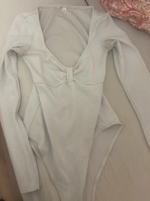 Vit ribbad långärmad body - Säljer en vit ribbad body från H&M med lång ärm och djup urringning. Bodyn har en snygg detalj framtill med en liten knut och är tillverkad i stretchigt material som sitter skönt mot kroppen. Perfekt att styla med jeans eller kjol.