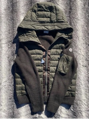 Grön pufferjacka från Moncler - Snygg grön pufferjacka från Moncler med stickade mörkbruna ärmar och rygg. Jackan har en stor huva, dragkedja framtill och en ficka med Moncler-logga på ena ärmen. Perfekt för kalla vinterdagar och riktigt trendig look.