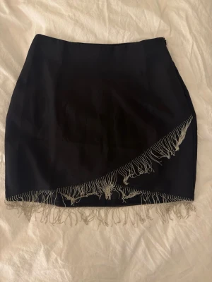 Svart kortkjol med fransdetalj - Aldrig använd svart kortkjol från Vero Moda med silvriga fransar längs nederkanten och sned detalj framtill. Perfekt till fest. Prislapp finns kvar. Köpt för 399kr.