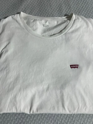 Vit t-shirt från Levi's med logga - Klassisk vit t-shirt från Levi's med rund halsringning och den ikoniska röda Levi's-loggan broderad på bröstet. Tillverkad i mjuk bomull för en skön känsla och enkel stil som funkar till allt.