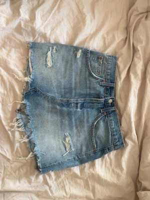 Blå sliten jeanskjol med fransar - Säljer en blå jeanskjol med råa fransade kanter och slitningar framtill och baktill. Kjolen är perfekt för sommaren och ger en avslappnad vibe.