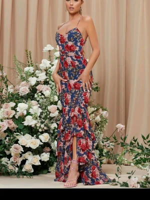 Blommig långklänning med volanger - Snygg långklänning med blå botten och stora röda och vita blommor. Klänningen har smala axelband som korsas i ryggen, volangdetaljer och en slits framtill. Perfekt för sommarens alla tillfällen.